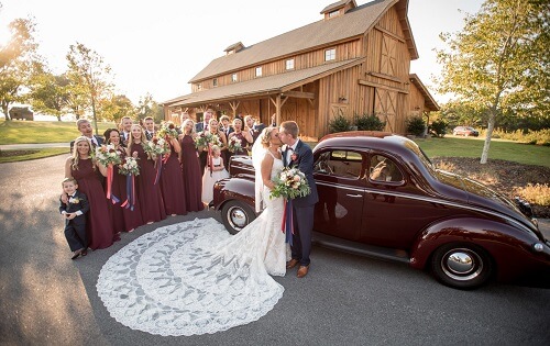 Bride and groom kissing next to antique car near bridal party standing in front of Windy Hill Wedding & Event Barn